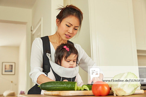 Frau mit kleiner Tochter im Tragetuch schneidet Salat an der Küchentheke