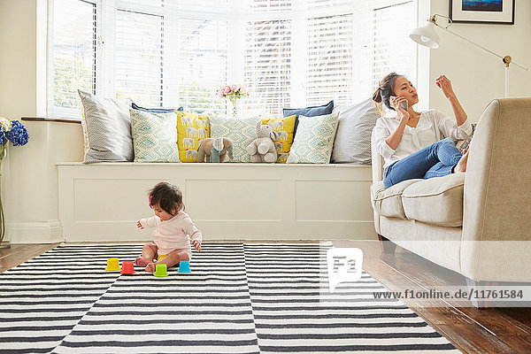 Frau mit Baby-Tochter auf Sofa liegend beim Smartphone-Anruf