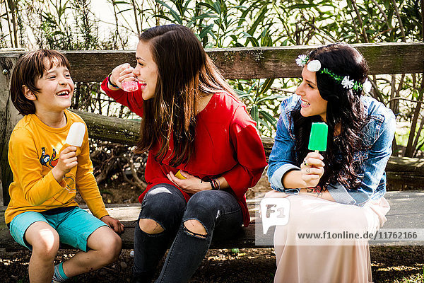 Zwei junge Frauen und ein kleiner Junge sitzen auf einer Bank  essen Eis am Stiel und lächeln