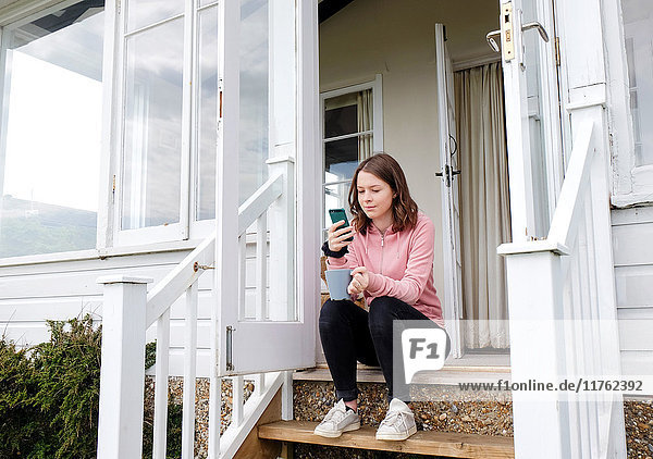 Junge Frau sitzt mit Kaffeetasse auf der Verandatreppe und schaut auf ein Smartphone