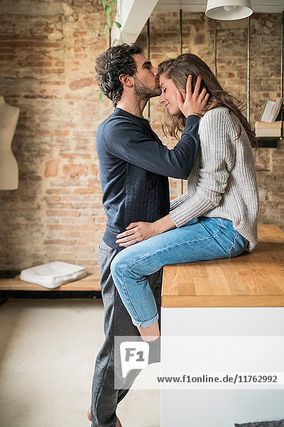 Junger Mann küsst Freundin auf der Küchenbank auf die Stirn
