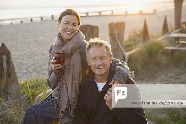 Portrait lächelndes reifes Paar mit Händchenhalten und Weintrinken am Strand bei Sonnenuntergang