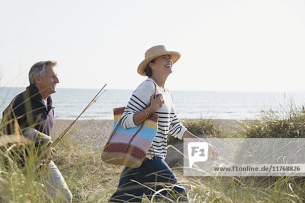 Mature couple with fishing rod walking in sunny beach grass