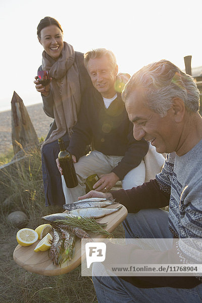 Reife Freunde trinken Wein und grillen Fische am Strand bei Sonnenuntergang.