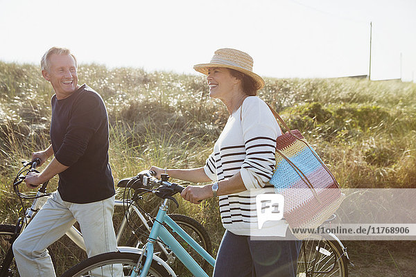 Mature couple walking bicycles on sunny beach grass path