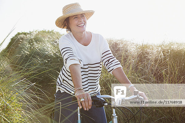 Smiling mature woman riding bicycle on sunny beach grass path