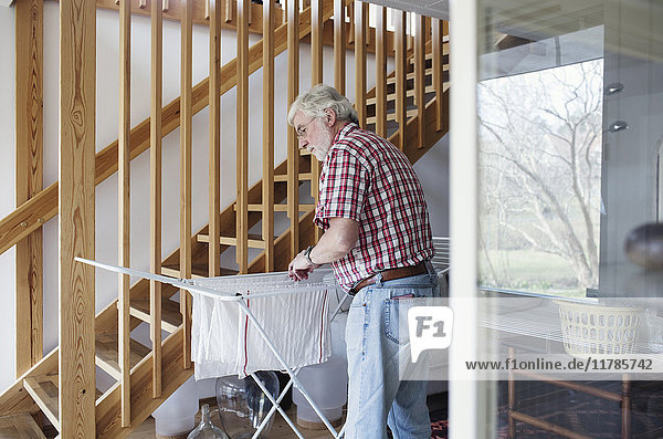 Senior Mann hängt Kleidung an einem Trockengestell  während er zu Hause an einer Holztreppe steht.
