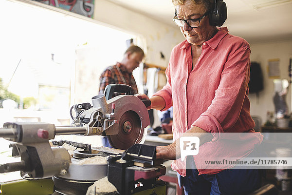 Senior woman using circular saw while man standing in background at workshop