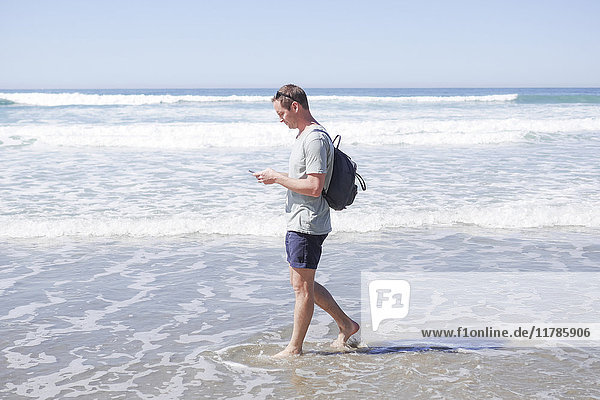 Full length side view of mature man using mobile phone while walking in sea at beach
