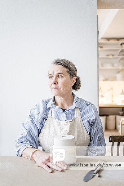 Senior female potter looking away while sitting at table with vase against white wall in workshop