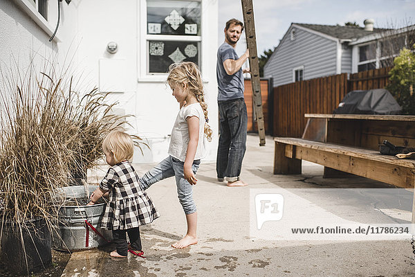 Vater mit Leiter beim Anblick von Töchtern  die außerhalb des Hauses mit Wasser spielen.