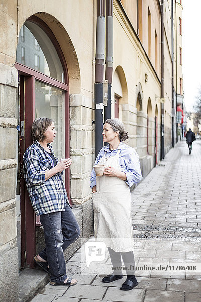 Fröhliche Kolleginnen stehen mit Kaffeetassen auf dem Fußweg beim Keramikladen in der Stadt