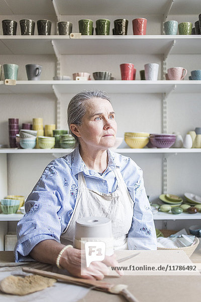 Senior female potter looking away while sitting at table with vase in workshop