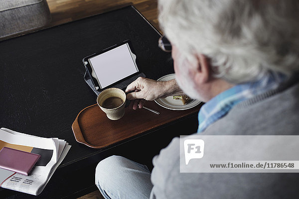 Abgeschnittenes Bild eines älteren Mannes  der eine Teetasse mit einem digitalen Tablett am Tisch hält.
