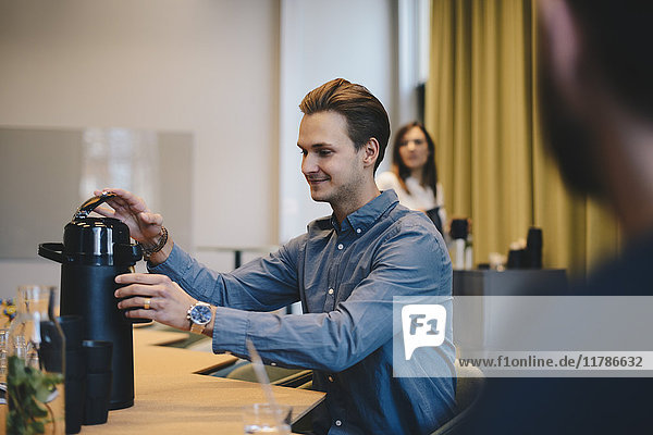 Lächelnder Geschäftsmann öffnet isolierten Getränkebehälter am Konferenztisch mit Kollegen im Sitzungssaal