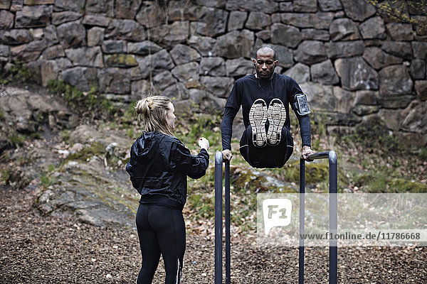 Rückansicht einer Sportlerin  die den Mann beim Training an parallelen Stäben gegen die Steinmauer im Wald beobachtet.