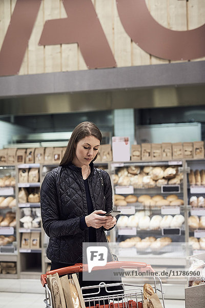 Frau mit Handy im Stehen mit Einkaufswagen am Regal im Supermarkt