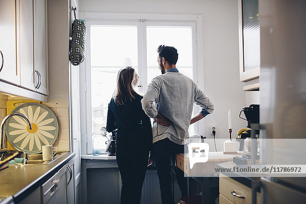 Rear view of couple talking while standing by dining table against window at home