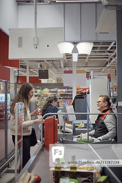 Frau im Gespräch mit reifer Kassiererin beim Bezahlen an der Kasse im Supermarkt