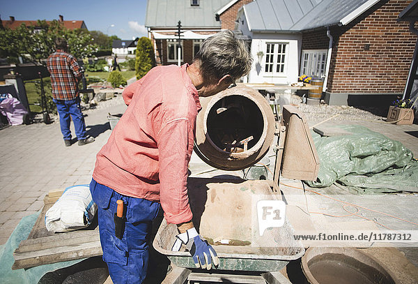 Side view of senior woman using cement mixer while man standing in background at yard