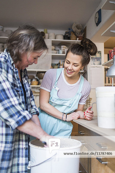 Happy woman looking at mature female potter mixing clay in bucket at store