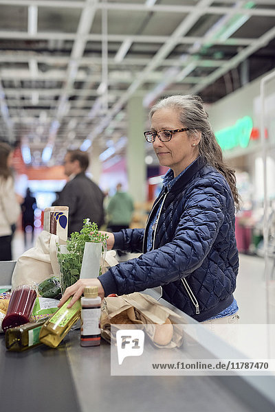 Reife Frau stehend mit Lebensmitteln und Tasche an der Kasse im Supermarkt