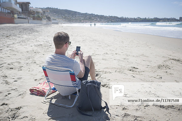 Rückansicht des Mannes  der ein Smartphone benutzt  während er auf einem Stuhl am Strand sitzt.