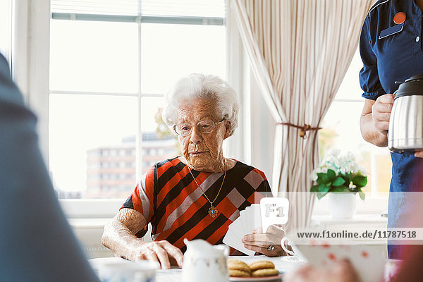 Seniorin beim Kartenspielen mit der Familie  während die Hausmeisterin die Kaffeekanne zu Hause hält.