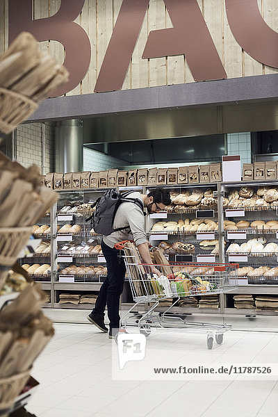 Mann hält Papiertüte im Einkaufswagen  während er im Supermarkt am Brotgestell steht.