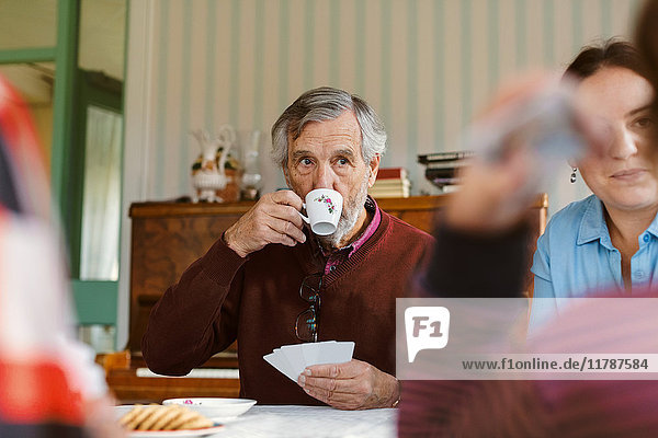 Senior man drinking coffee while playing cards with family at home