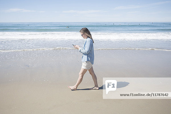 Full length side view of woman using mobile phone while walking at beach