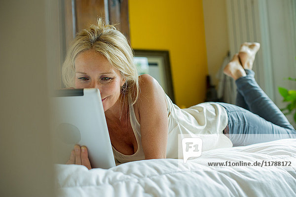 Reife Frau  die auf dem Bett liegt und ein digitales Tablett sieht.