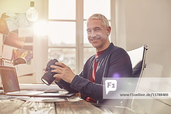 Portrait lächelnder  selbstbewusster Fotograf mit Digitalkamera bei der Arbeit am Laptop im sonnigen Büro