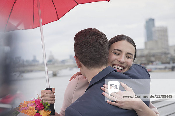 Smiling  affectionate couple with umbrella and flowers hugging on urban bridge  London  UK