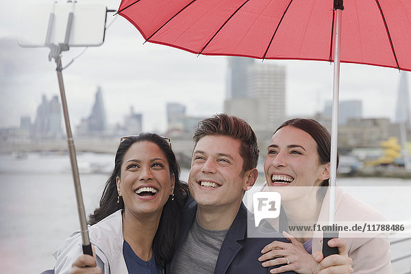 Smiling friend tourists with umbrella taking selfie with selfie stick  London  UK