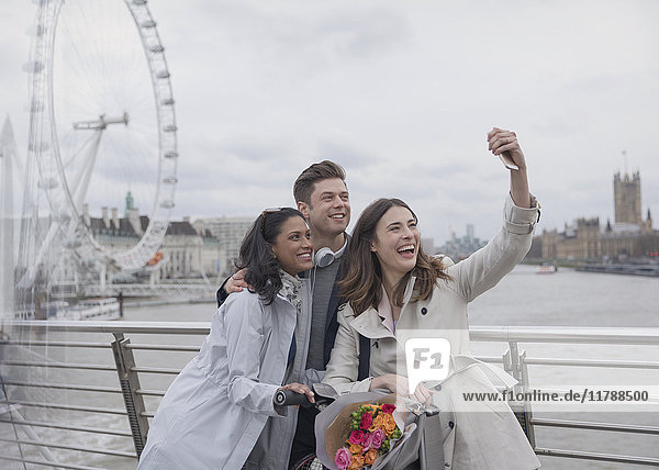 Smiling  happy friends taking selfie with selfie stick on bridge near Millennium Wheel  London  UK
