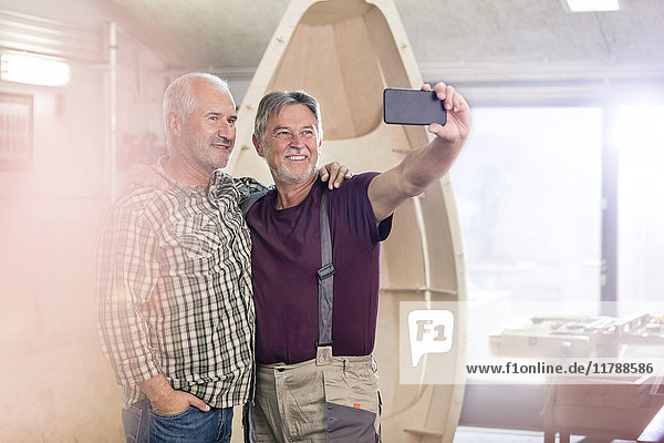 Stolze  lächelnde Schreiner mit Fotohandy  die Selfie neben dem Holzboot in die Werkstatt bringen.