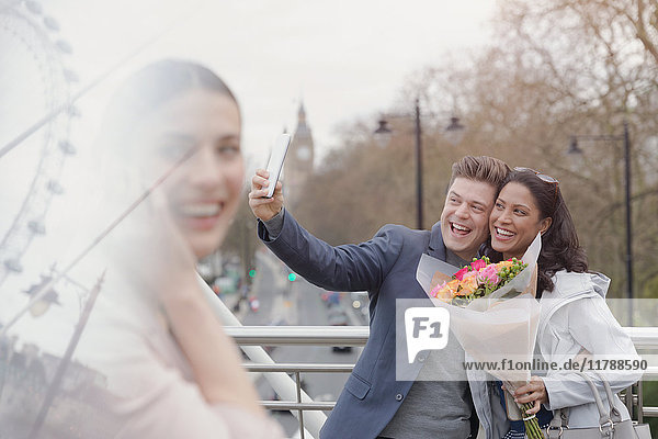 Enthusiastisches Paar mit Blumenstrauß  das Selfie mit Fotohandy auf urbaner Brücke nimmt  London  UK