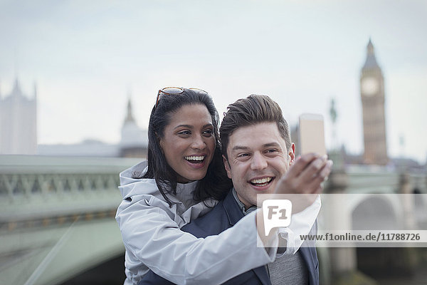 Playful  affectionate couple tourists taking selfie with camera phone in front of Westminster Bridge  London  UK