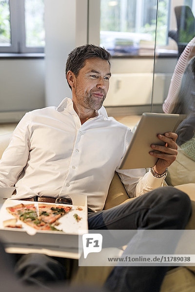 Businessman in bean bag in office eating pizza and using tablet