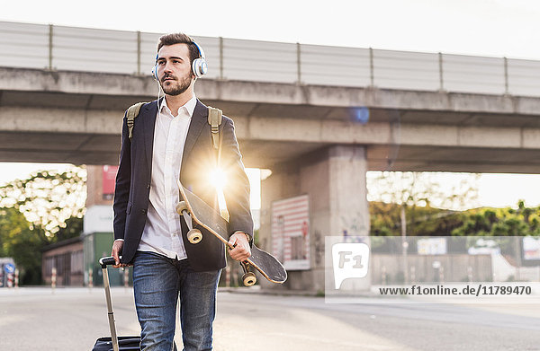 Young man on the move with skateboard  rolling suitcase and headphones