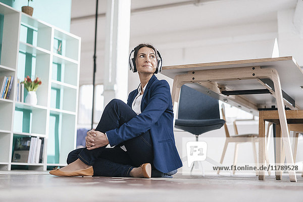 Geschäftsfrau sitzt auf dem Boden in einem Loft und hört Musik mit Kopfhörern.