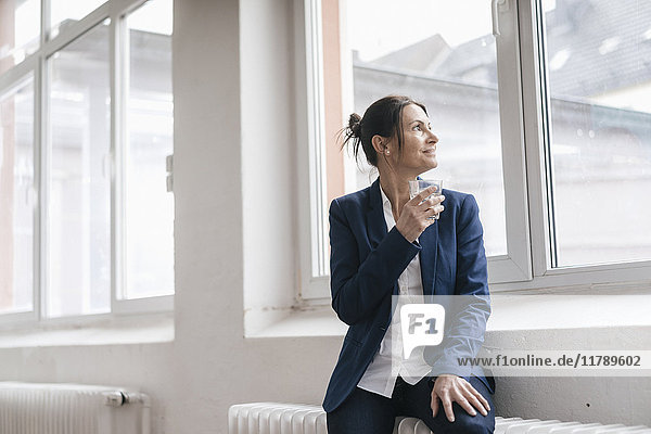 Businesswoman with glass of water sitting on radiator in a loft looking through window