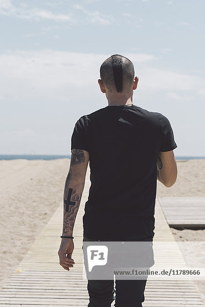 Rückansicht eines jungen Mannes mit Irokesenschnitt und Tattoos  der auf einer Strandpromenade zum Strand geht.