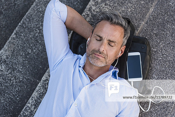 Mann mit geschlossenen Augen auf der Treppe liegend mit Handy und Ohrstöpseln