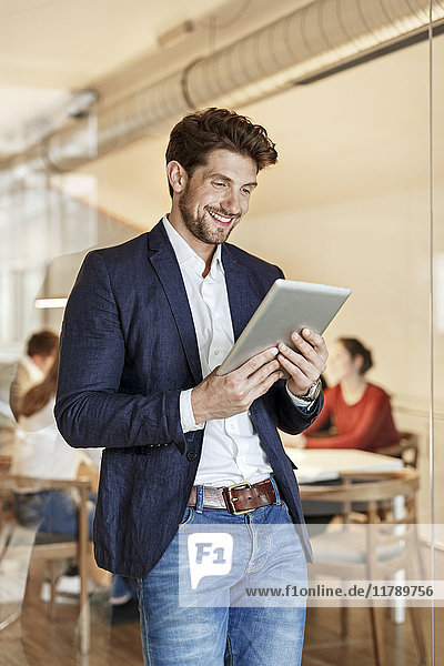 Smiling businessman using tablet in office with a meeting in background