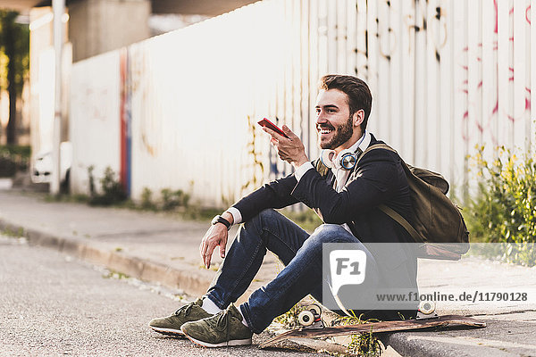 Smiling young man sitting on pavement using cell phone