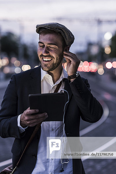 Happy young man in the city with earphones and tablet at dusk