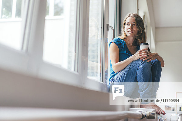 Lächelnde junge Frau mit Kaffee zum Mitnehmen am Fenster sitzend