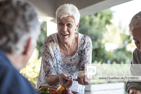 Porträt einer lächelnden Seniorin  die mit Hilfe ihrer Freunde das Mittagessen auf der Terrasse zubereitet.